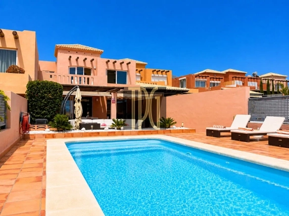 Sunlit resort pool area with terracotta deck, pink-beige buildings, and white lounge chairs under a clear blue sky.