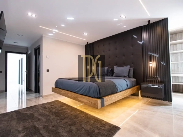 Contemporary bedroom with a low wooden platform bed, a tufted black headboard wall, gray bedding, and a glossy black nightstand. A large dark rug sits on a light tiled floor, with hallway access visible to the left.