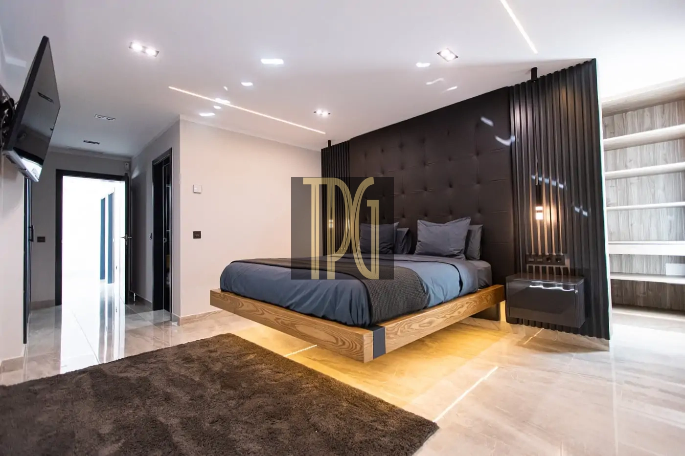 Contemporary bedroom with a low wooden platform bed, a tufted black headboard wall, gray bedding, and a glossy black nightstand. A large dark rug sits on a light tiled floor, with hallway access visible to the left.