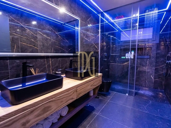 Luxurious bathroom with black marble walls, blue LED lighting, and a wooden vanity with a black vessel sink A modern bathroom featuring a glass-walled shower, dark marble surfaces, and neon blue accent lighting Bathroom with a wide mirror, wooden counter, black basin sink, rolled towels on the shelf, and a rainfall shower in a glass enclosure Contemporary spa-like bathroom showing a illuminated ceiling, dark stone tiles, and a spacious glass shower area Black marble bathroom with blue LED lines, floating wooden vanity, and a large rainfall shower door system