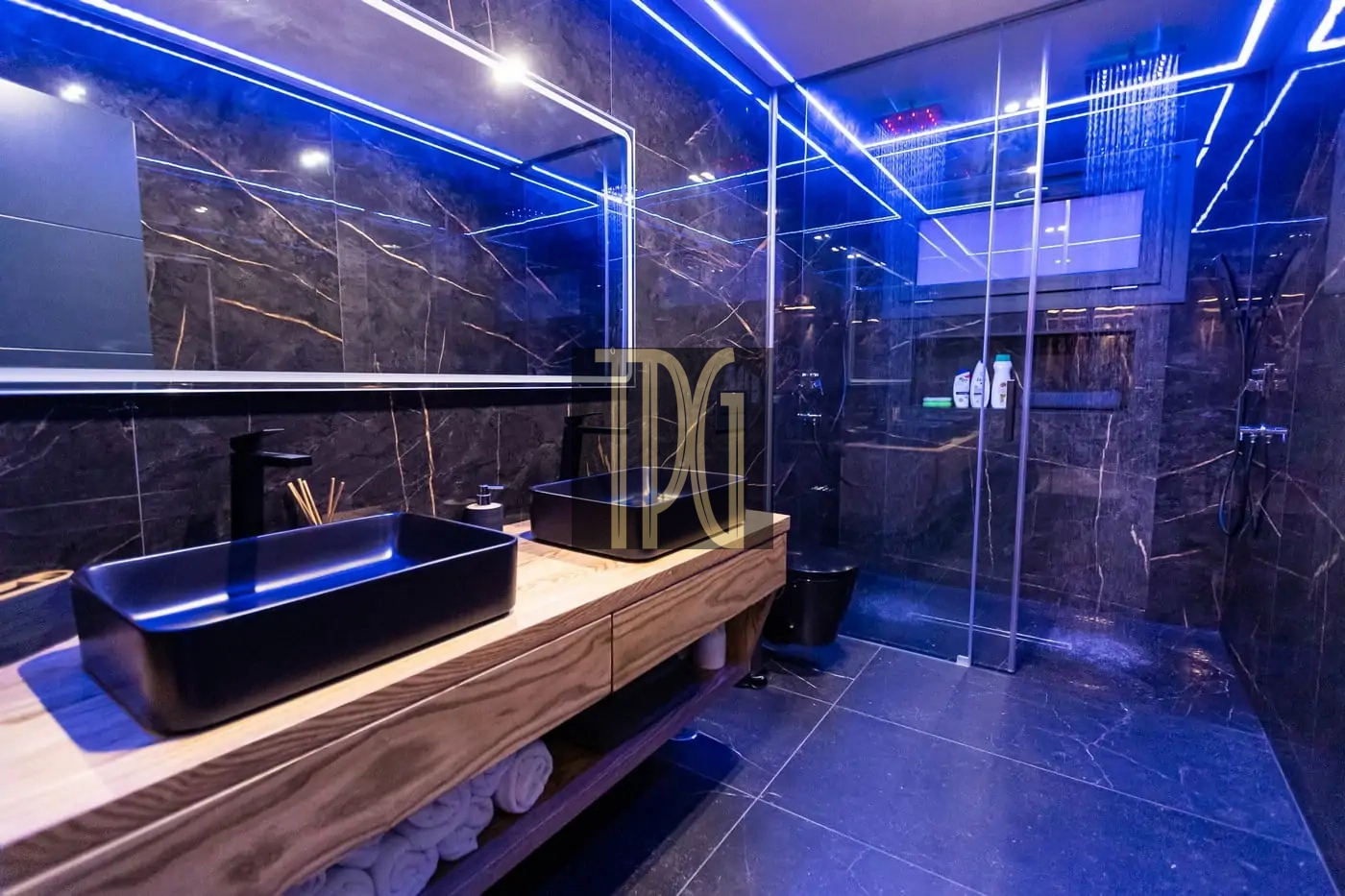 Luxurious bathroom with black marble walls, blue LED lighting, and a wooden vanity with a black vessel sink A modern bathroom featuring a glass-walled shower, dark marble surfaces, and neon blue accent lighting Bathroom with a wide mirror, wooden counter, black basin sink, rolled towels on the shelf, and a rainfall shower in a glass enclosure Contemporary spa-like bathroom showing a illuminated ceiling, dark stone tiles, and a spacious glass shower area Black marble bathroom with blue LED lines, floating wooden vanity, and a large rainfall shower door system