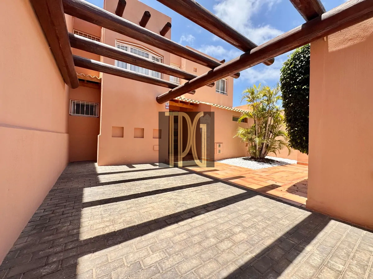 File 00010jpg Outdoor courtyard with peach walls, brick pavers, and overhead wooden pergola beams casting shadows under a bright blue sky.