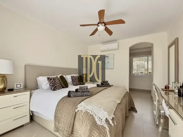 Bright beige master bedroom with a king bed, leopard-print pillows, folded towels, and a ceiling fan above.