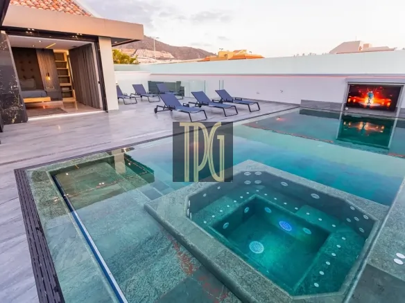 Luxurious rooftop pool area with a glass-edged pool, a built-in hot tub, and blue lounge chairs on a marble deck, plus an open bedroom entry in the background and distant hills.