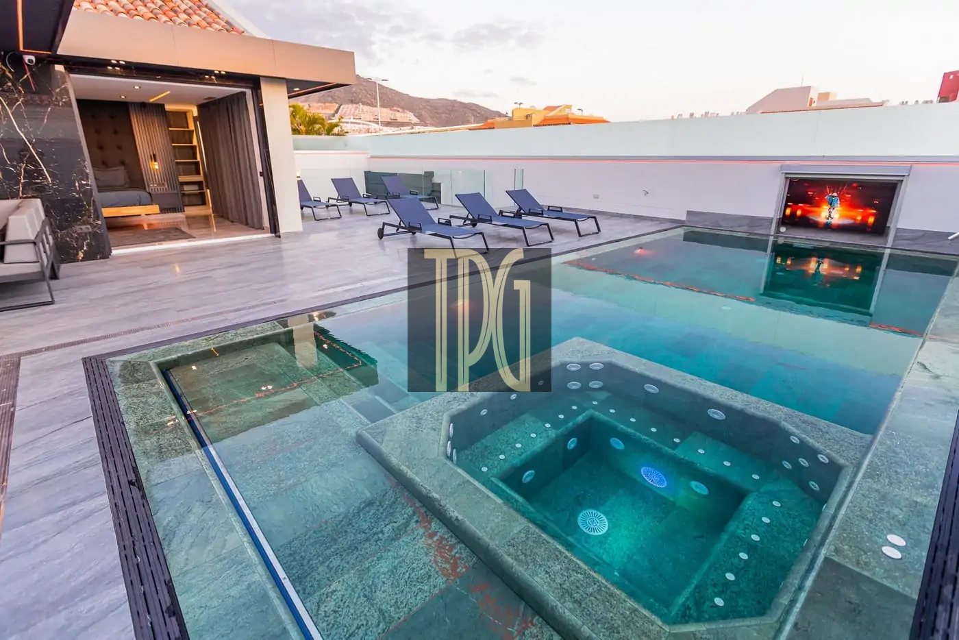 Luxurious rooftop pool area with a glass-edged pool, a built-in hot tub, and blue lounge chairs on a marble deck, plus an open bedroom entry in the background and distant hills.