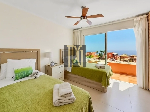 Hotel room with two single beds in green bedspreads, a ceiling fan, and a balcony overlooking orange rooftops and the ocean; towels folded on each bed; soft white curtains and a bedside table with lamp and decor.
