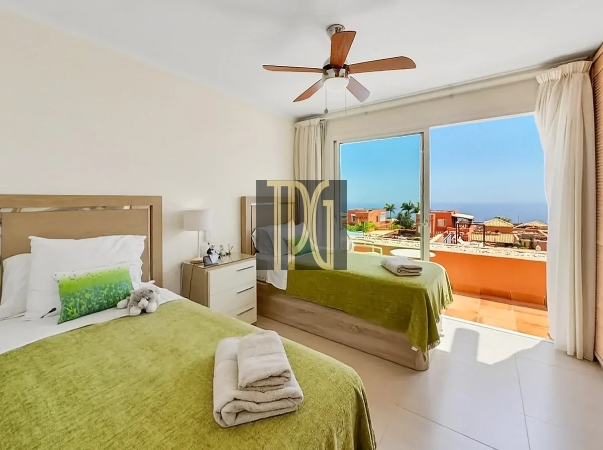 File 00013jpg Hotel room with two single beds in green bedspreads, a ceiling fan, and a balcony overlooking orange rooftops and the ocean; towels folded on each bed; soft white curtains and a bedside table with lamp and decor.
