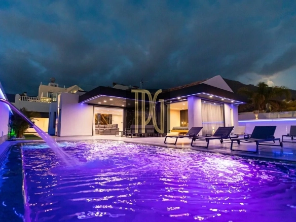 Modern white villa with a purple-lit swimming pool and lined-up lounge chairs at night.