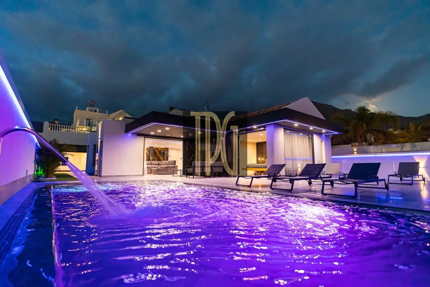 Modern white villa with a purple-lit swimming pool and lined-up lounge chairs at night.