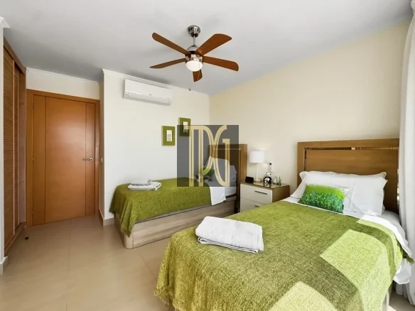 Hotel room with two single beds, green bedspreads, and white pillows; wooden headboards, nightstand, and a lamp between the beds. A ceiling fan and wall-mounted AC unit above, plus a wardrobe on the left.