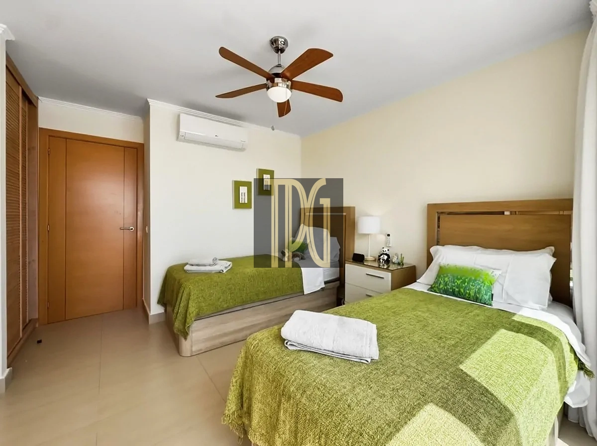 File 00014jpg Hotel room with two single beds, green bedspreads, and white pillows; wooden headboards, nightstand, and a lamp between the beds. A ceiling fan and wall-mounted AC unit above, plus a wardrobe on the left.