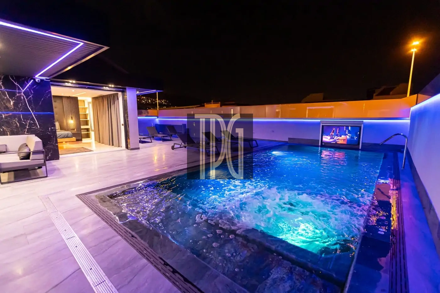 Rooftop pool at night with blue lighting, lounge chairs, and an open sliding-door living area visible to the left.