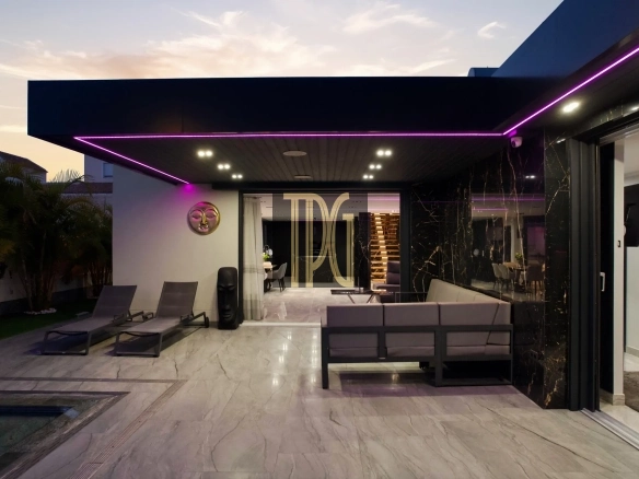 Modern outdoor patio at dusk with purple LED trim, lounge chairs, and a glass-walled living area inside.
