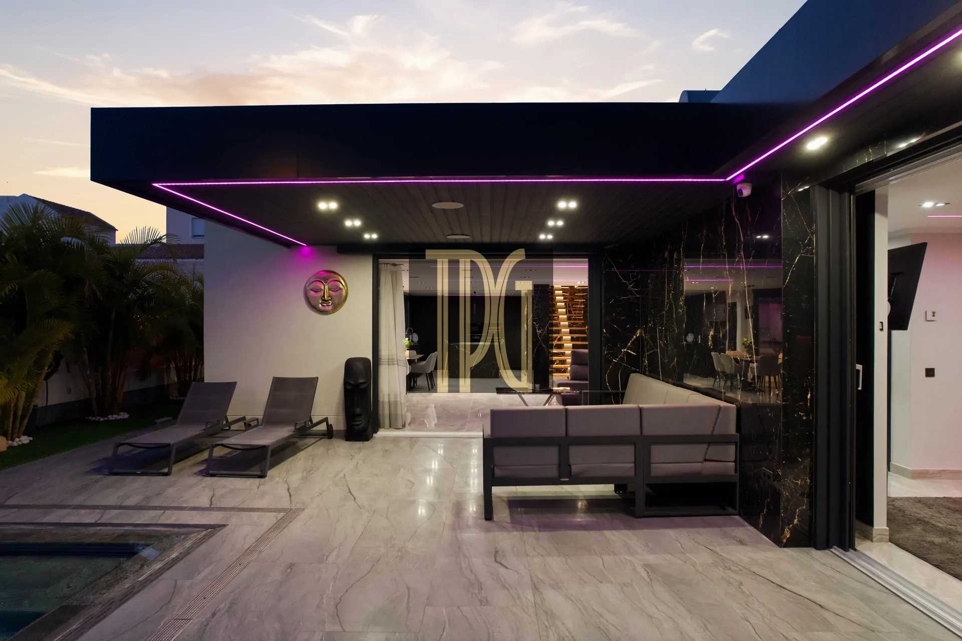 Modern outdoor patio at dusk with purple LED trim, lounge chairs, and a glass-walled living area inside.