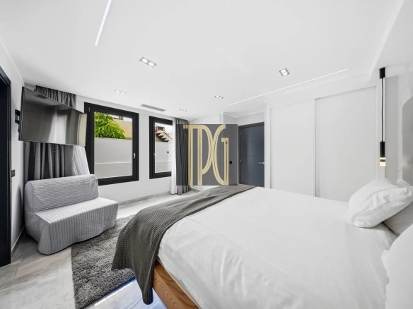 Modern white hotel bedroom with two beds, gray bedding, a wall-mounted TV, and large windows with black frames.",