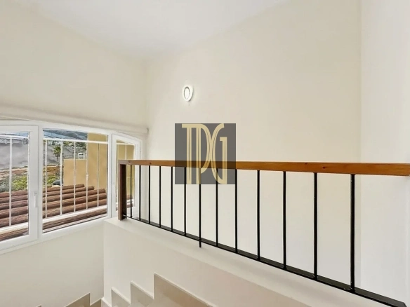 Indoor stairwell with white walls, a wooden handrail, and black metal balusters, next to a large window with bars visible outside.