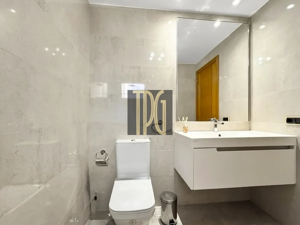 File 00020jpg Modern beige-toned bathroom with a wall-mounted white vanity and rectangular sink, large mirror, and a white toilet beneath a tiled wall. A small trash can sits on the floor and a toothbrush holder is on the counter.