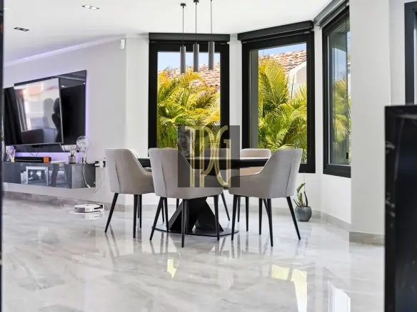 Contemporary dining room with a glass-top table, six gray chairs, and large windows with tropical plants outside.