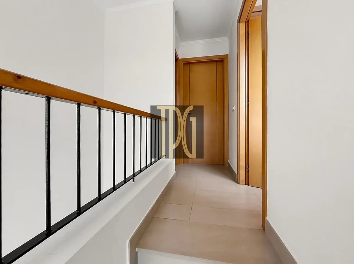File 00021jpg Bright upstairs hallway with a wooden handrail and black metal balusters, beige tiled floor, and closed wooden doors along the right edge.