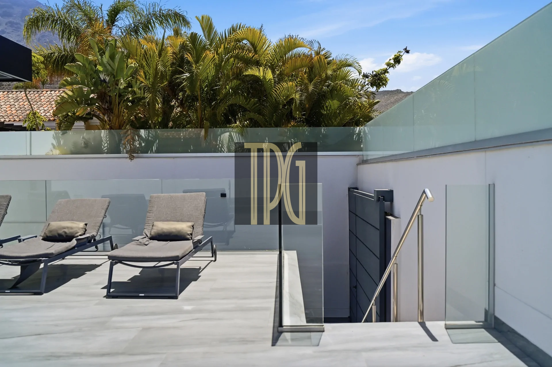 Modern rooftop terrace with two gray lounge chairs, glass railing, and palm trees against a clear blue sky.