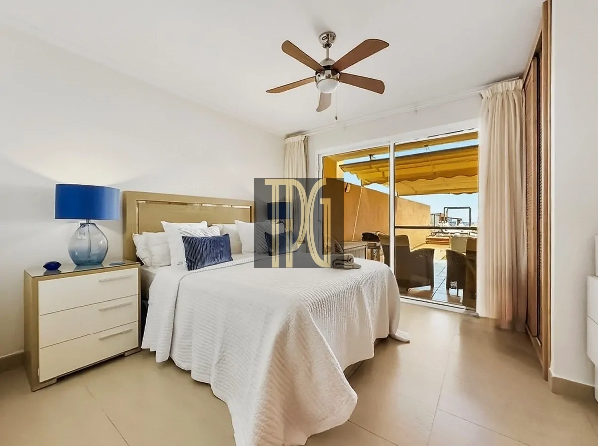 File 00024jpg Bright bedroom with white bedding, beige headboard, blue glass lamp on a nightstand, and a balcony door leading to outdoor seating.