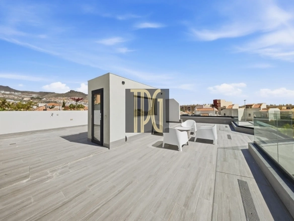 Rooftop terrace with light-gray tiled floor, white cubical structure, glass railing, and white plastic chairs under a blue sky with a distant hillside town in the background