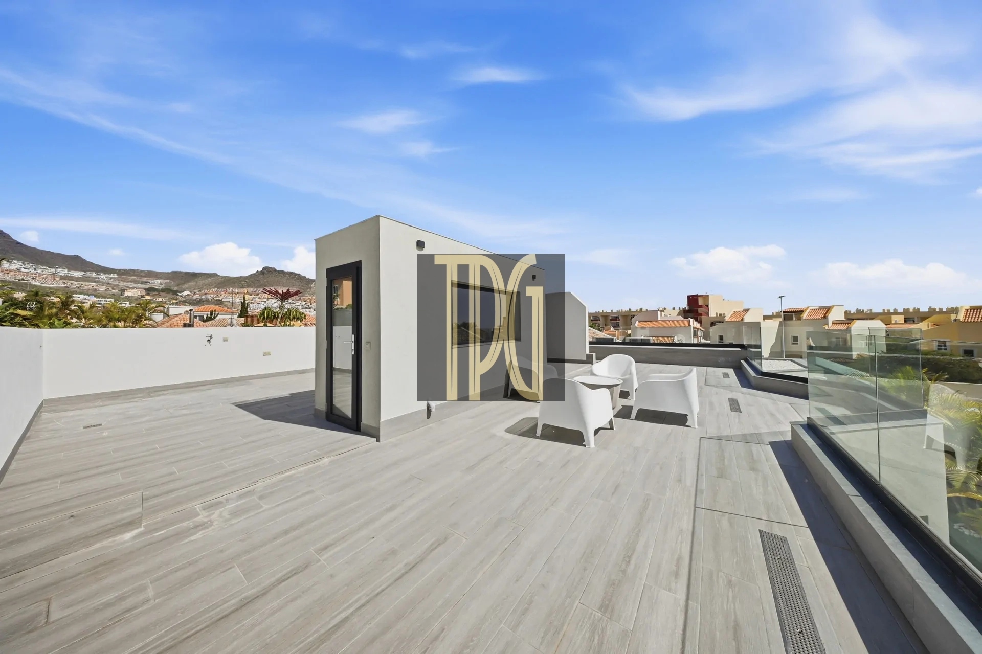 Rooftop terrace with light-gray tiled floor, white cubical structure, glass railing, and white plastic chairs under a blue sky with a distant hillside town in the background