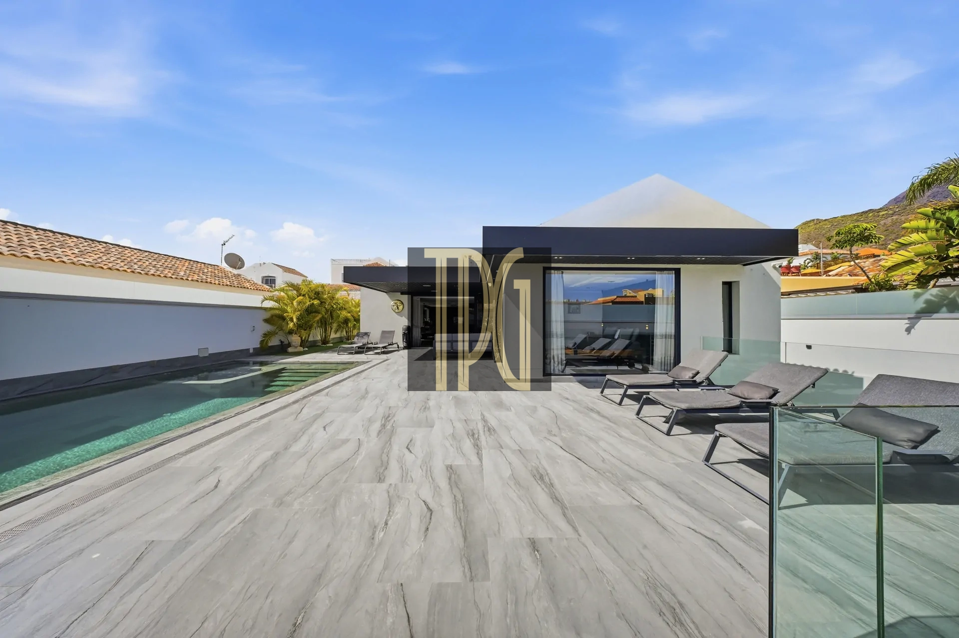 Modern villa backyard with a rectangular pool, lounge chairs, glass railing, and a blue sky above.