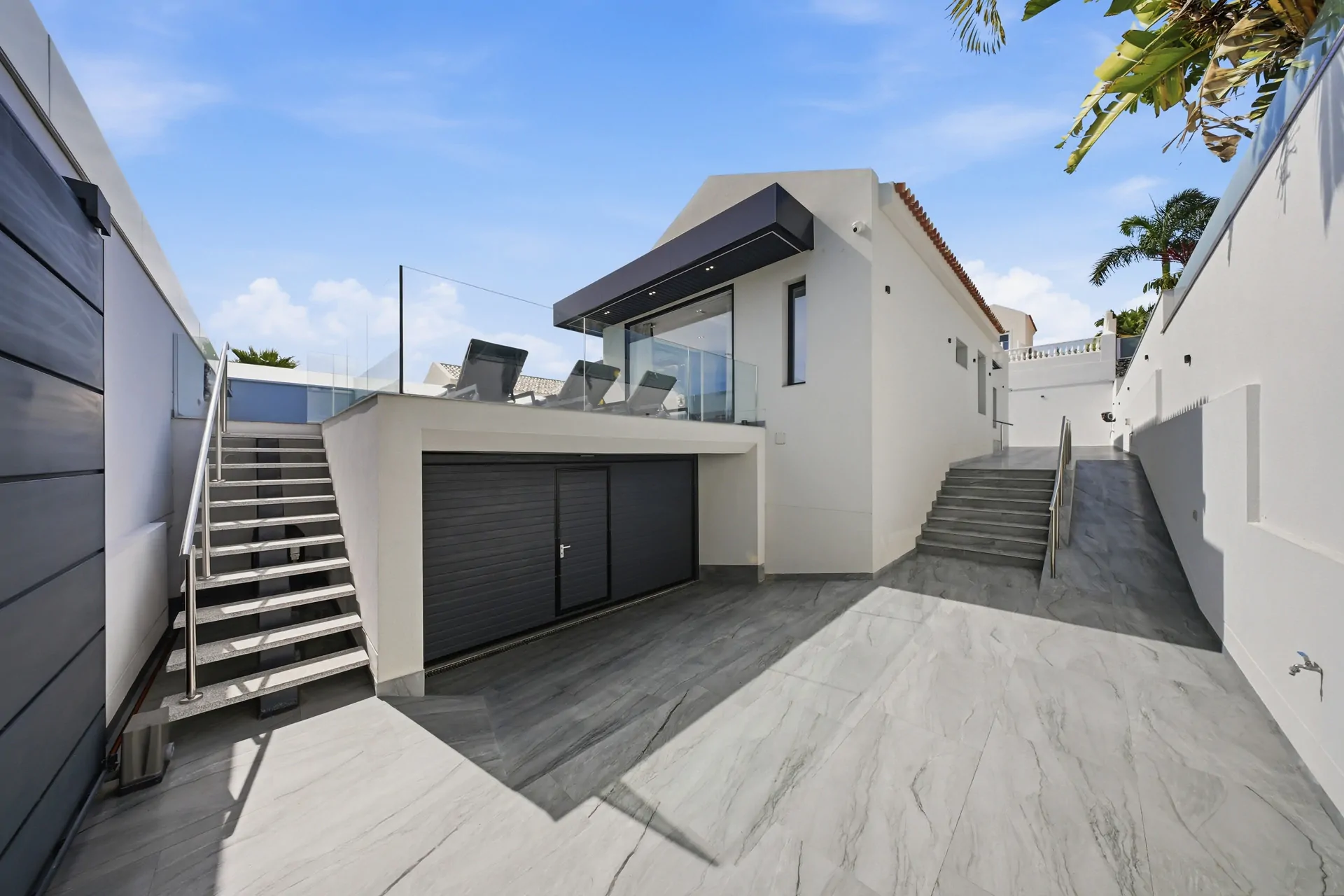 Modern white contemporary home with glass-railed balcony, gray garage doors, and marble courtyard under a blue sky.
