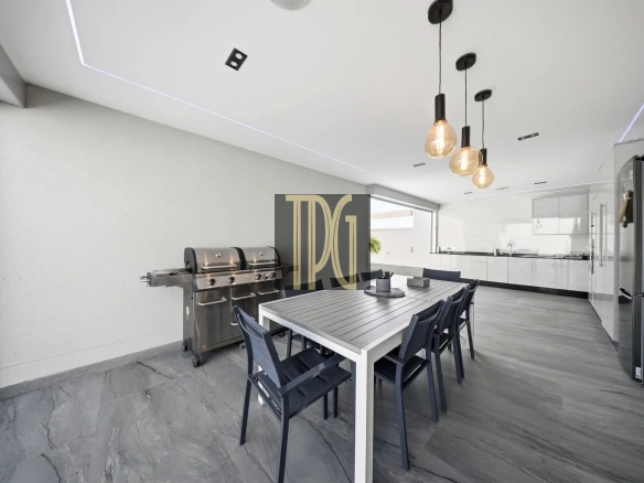 Modern outdoor dining area with a long white table, dark chairs, and three hanging Edison bulbs; stainless grill on the left and a sleek kitchen in the background.