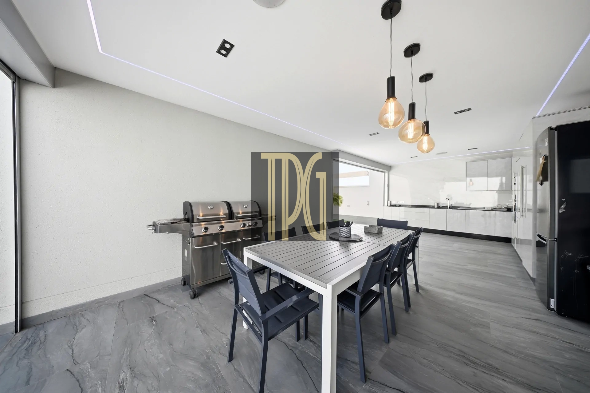 Modern outdoor dining area with a long white table, dark chairs, and three hanging Edison bulbs; stainless grill on the left and a sleek kitchen in the background.