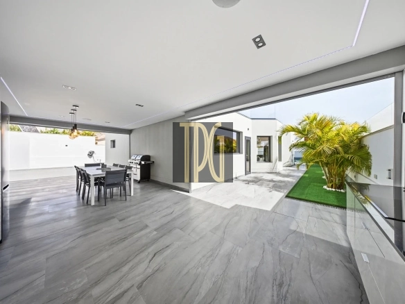 Open-plan indoor-outdoor living space with a gray marble floor, dining table, and a barbecue grill, opening to a sunny courtyard with palm trees on artificial turf.