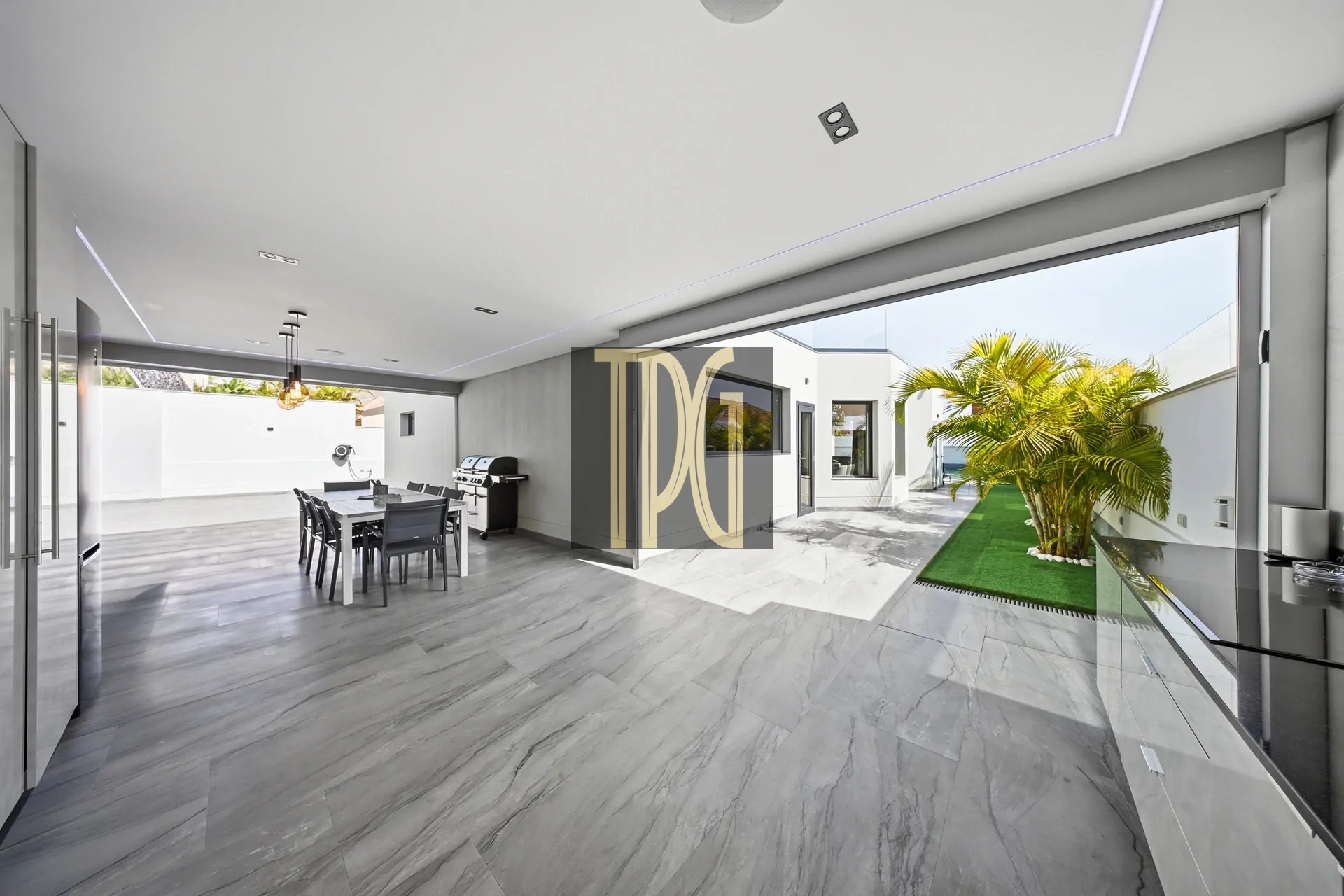 Open-plan indoor-outdoor living space with a gray marble floor, dining table, and a barbecue grill, opening to a sunny courtyard with palm trees on artificial turf.