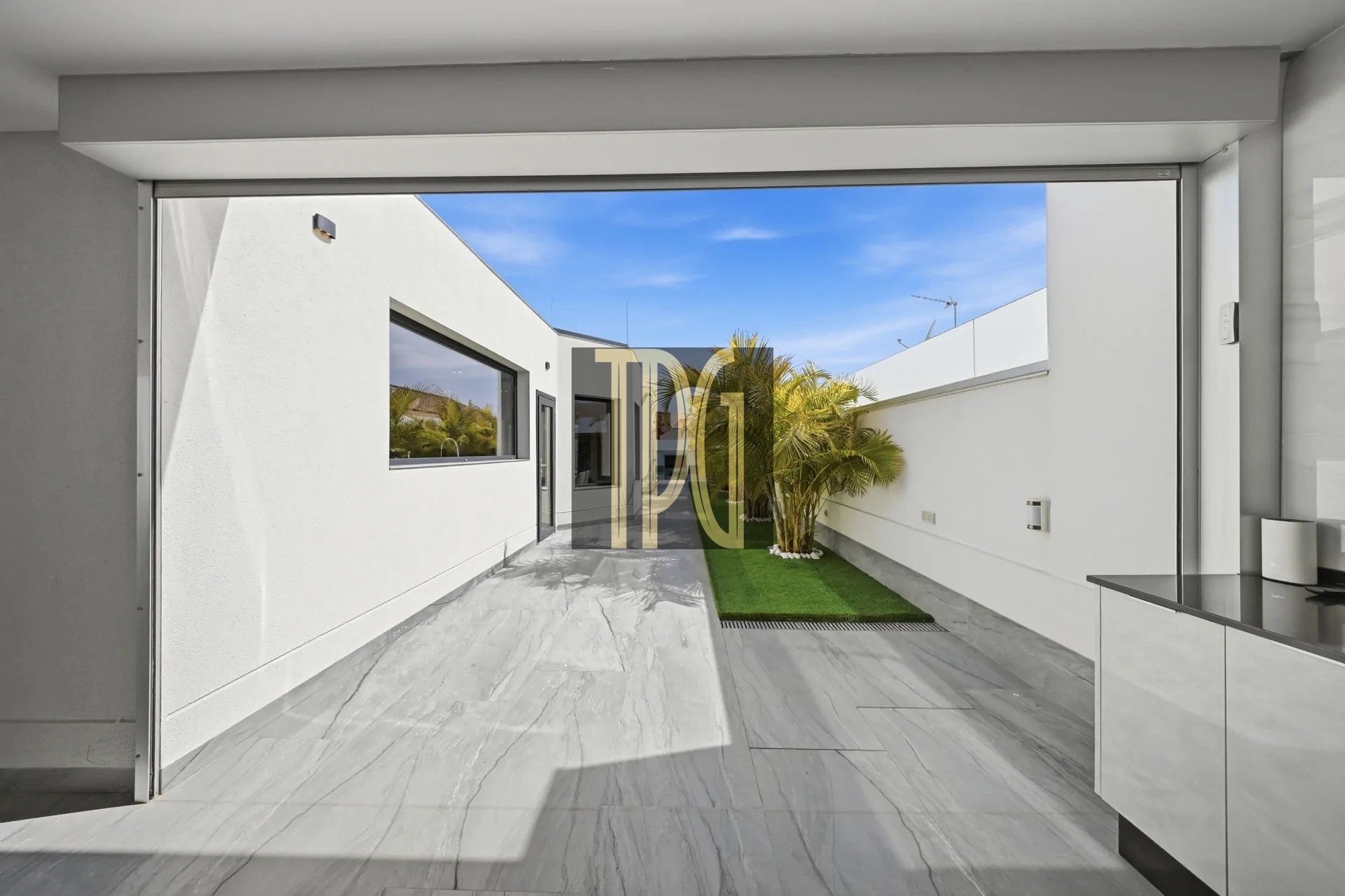 Interior view of a modern kitchen opening to a white courtyard with a palm tree, green lawn, and blue sky beyond.