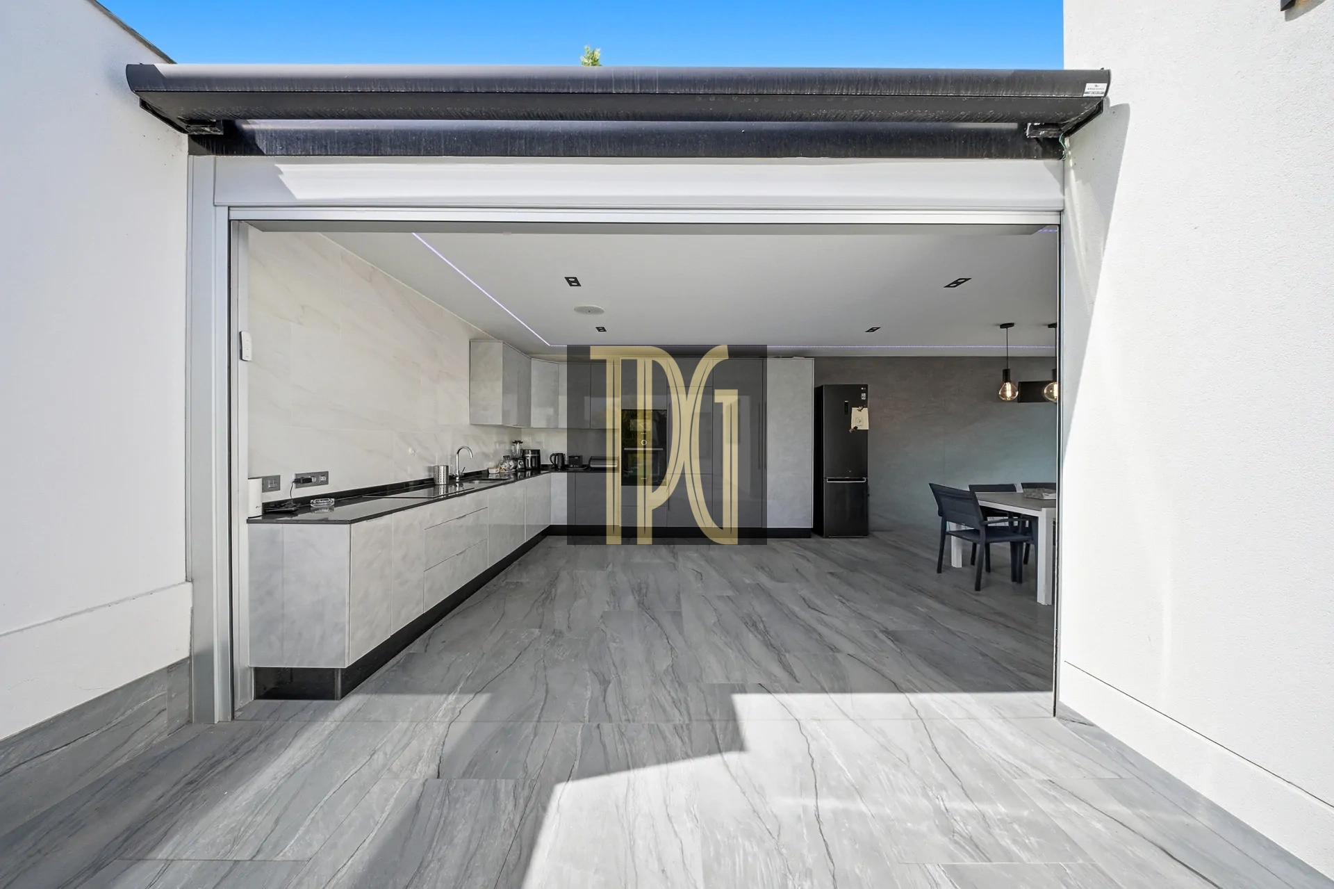 Open garage door reveals a modern, open-concept kitchen with white cabinets, gray marble floors, and a dining area in the back.