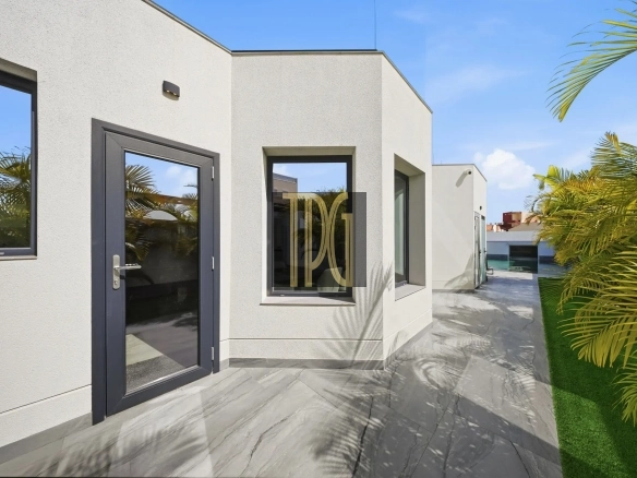 Modern white stucco building with a dark-framed glass door and window, on a tiled patio under a bright blue sky, with palm plants nearby.