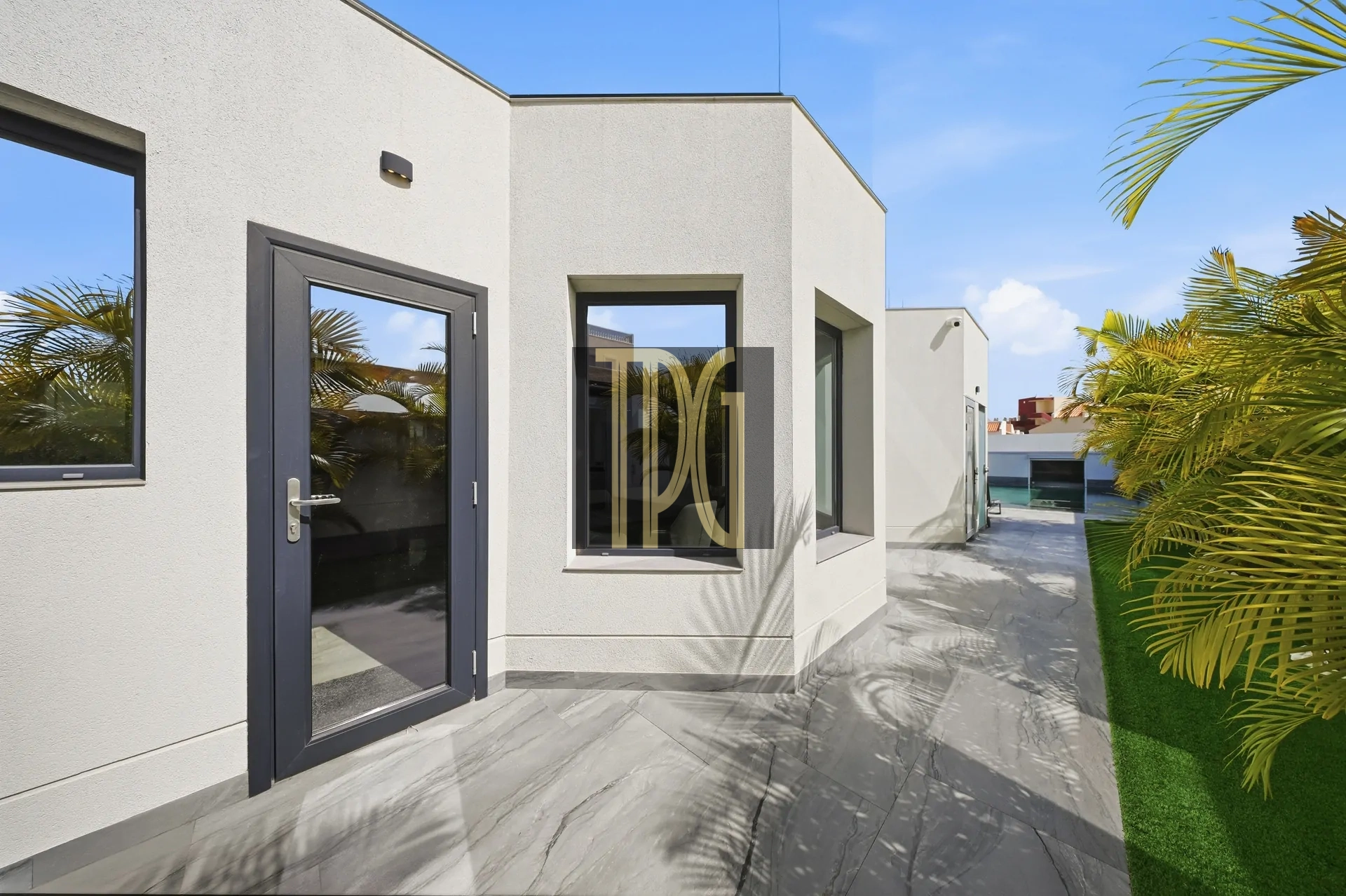 Modern white stucco building with a dark-framed glass door and window, on a tiled patio under a bright blue sky, with palm plants nearby.