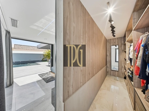 Walk-in closet with wooden panels, hanging clothes on the right, and a glass door opening to a sunny outdoor space with a pool.