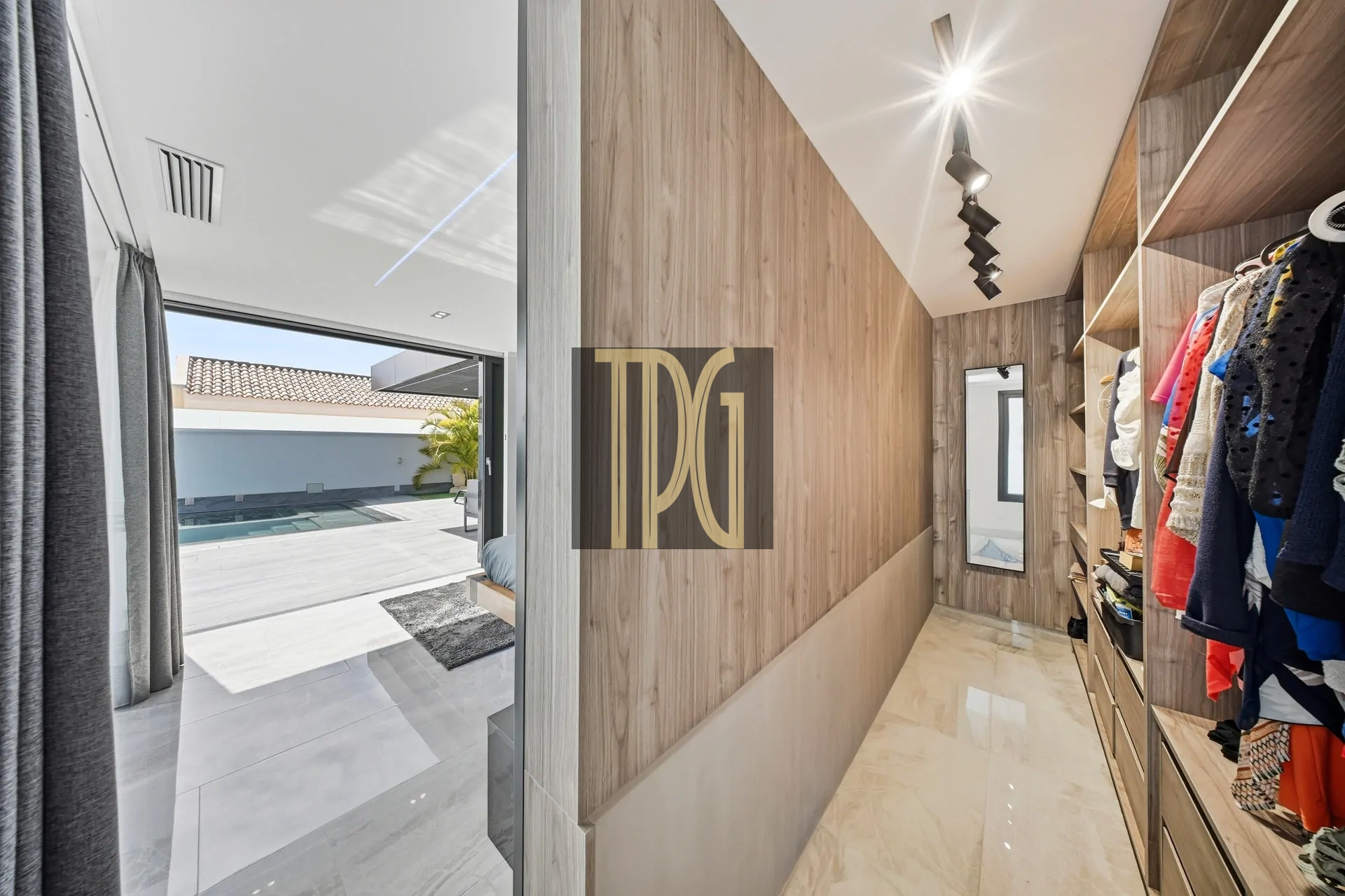 Walk-in closet with wooden panels, hanging clothes on the right, and a glass door opening to a sunny outdoor space with a pool.