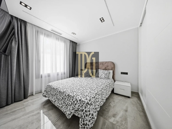 Modern bedroom with a double bed, gray patterned duvet, and white nightstand under a large window with curtains. A wall-mounted TV is angled near the head of the bed.