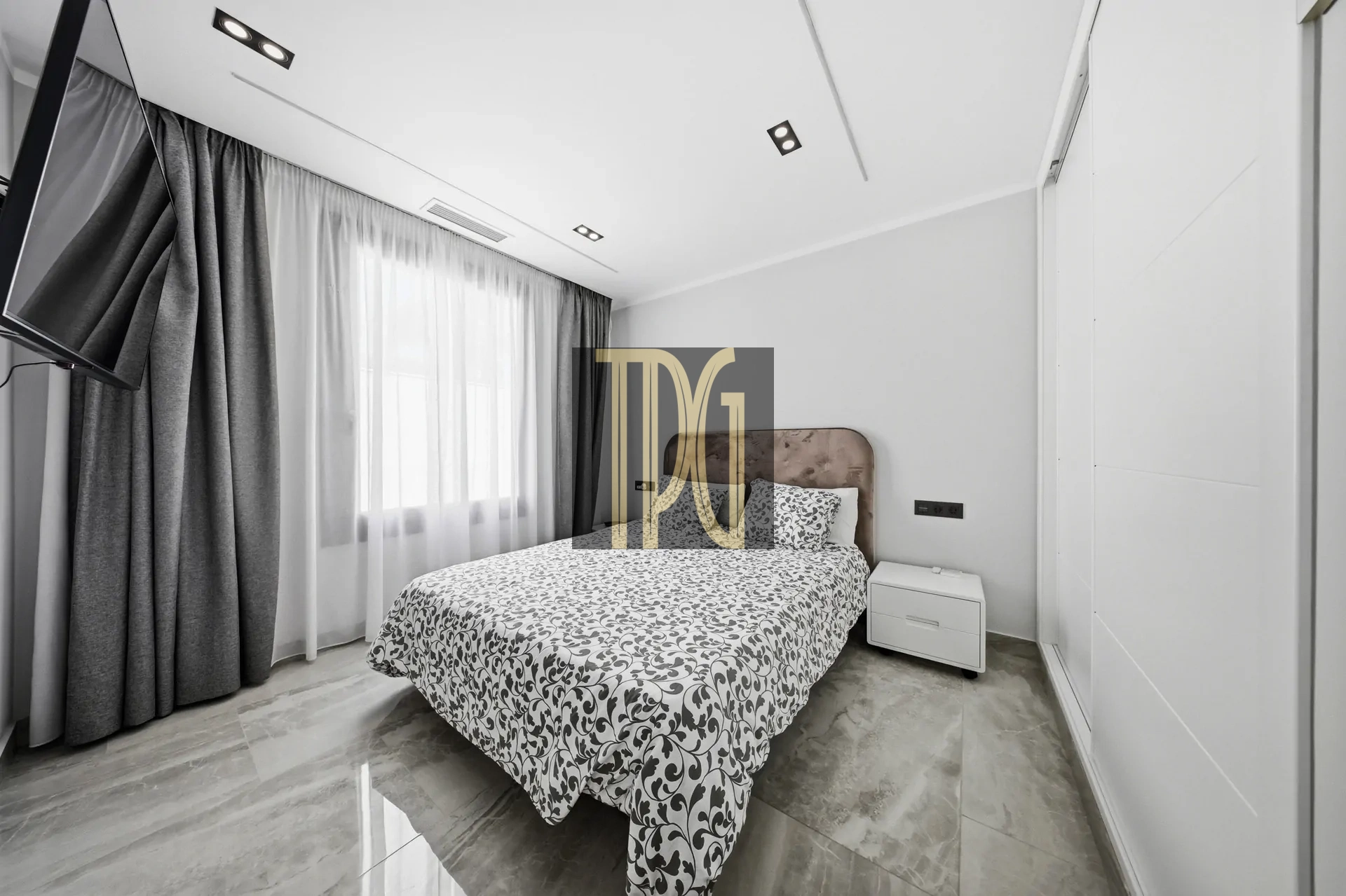 Modern bedroom with a double bed, gray patterned duvet, and white nightstand under a large window with curtains. A wall-mounted TV is angled near the head of the bed.