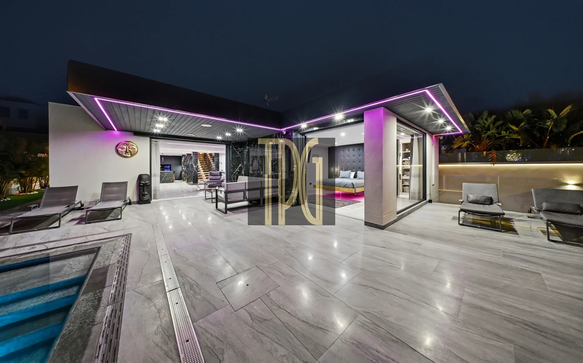 Night view of a modern backyard with a glass-walled living area, purple LED accents along the roof, and lounge chairs on a tiled patio.