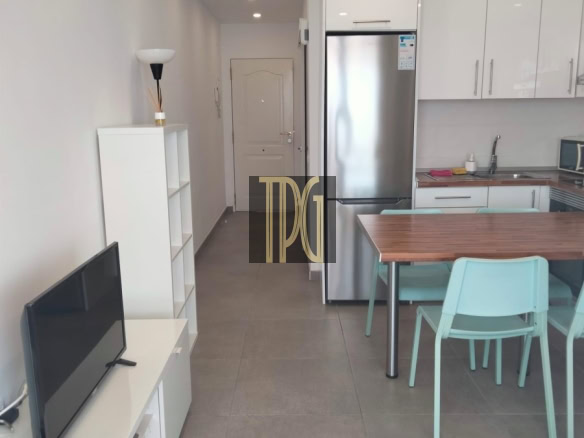 Open-plan living area with a white TV stand and flat-screen TV on the left, a tall white shelf, and a stainless steel fridge in a modern kitchen; wooden dining table with teal chairs to the right.