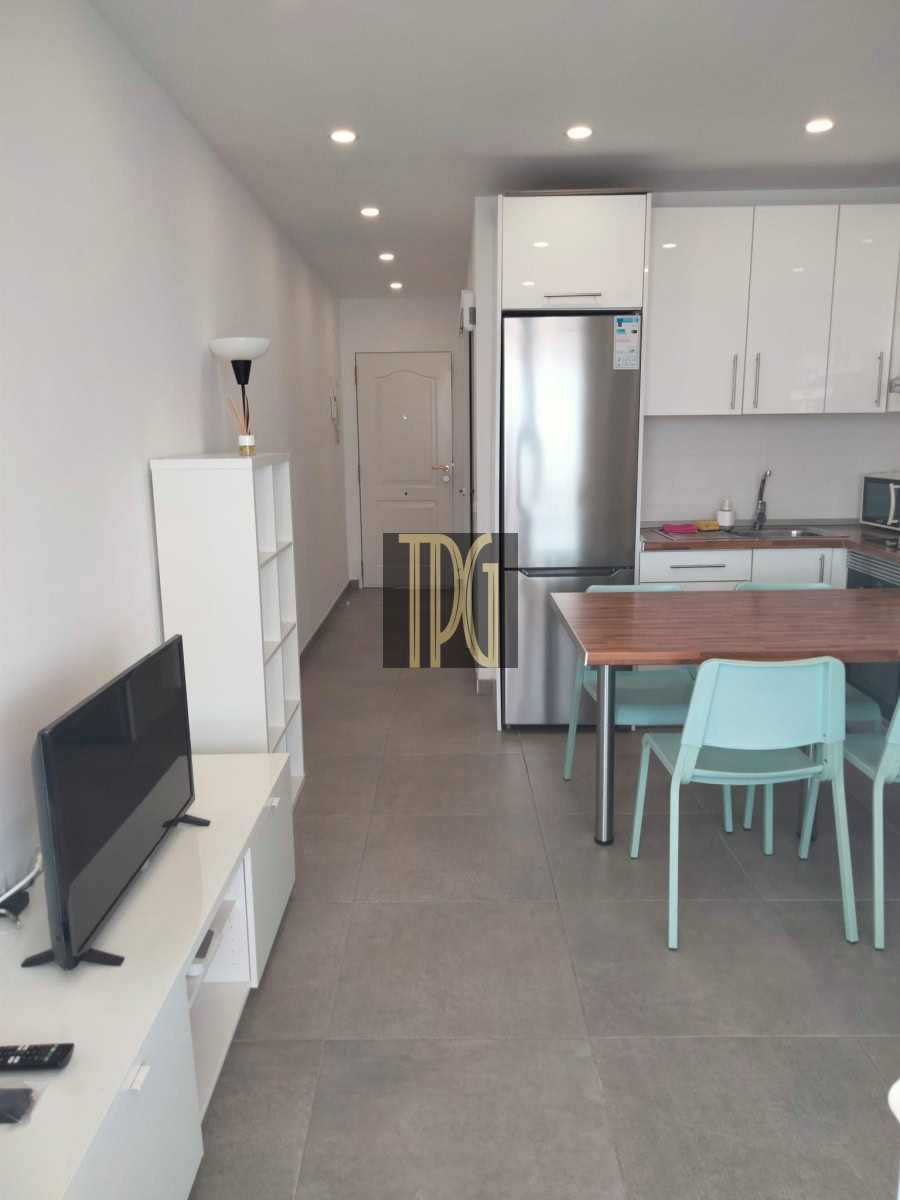 Open-plan living area with a white TV stand and flat-screen TV on the left, a tall white shelf, and a stainless steel fridge in a modern kitchen; wooden dining table with teal chairs to the right.