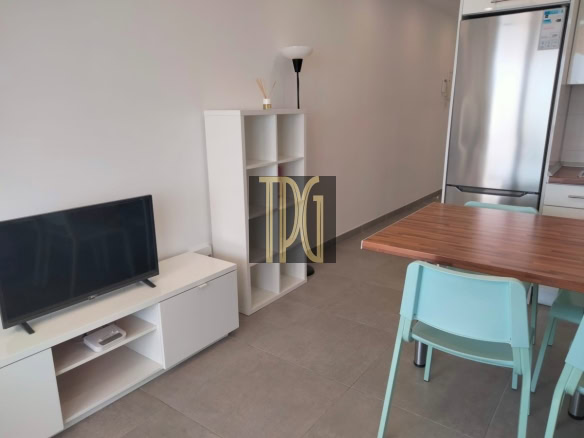 Modern open-plan living area: TV on a white unit, cubby shelf, floor lamp, wooden dining table, and mint-green chairs with a stainless fridge in the background.