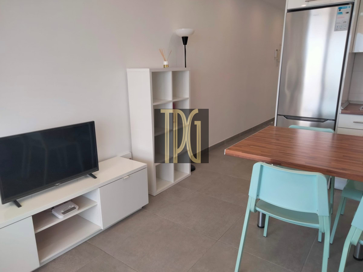 Modern open-plan living area: TV on a white unit, cubby shelf, floor lamp, wooden dining table, and mint-green chairs with a stainless fridge in the background.