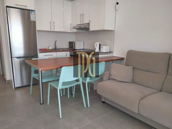 Open-plan kitchen and living area with a wooden dining table and light turquoise chairs beside a beige sofa. Stainless steel refrigerator next to white upper cabinets, with a microwave, kettle, and toaster on the counter. Turquoise dining chairs surround a wooden table in a modern kitchen setup with a gray-tiled floor. Beige sofa in a cozy living space adjacent to a compact, contemporary kitchen in a studio apartment. Modern apartment: kitchen and living area with white cabinets, stainless appliances, and a small dining table.
