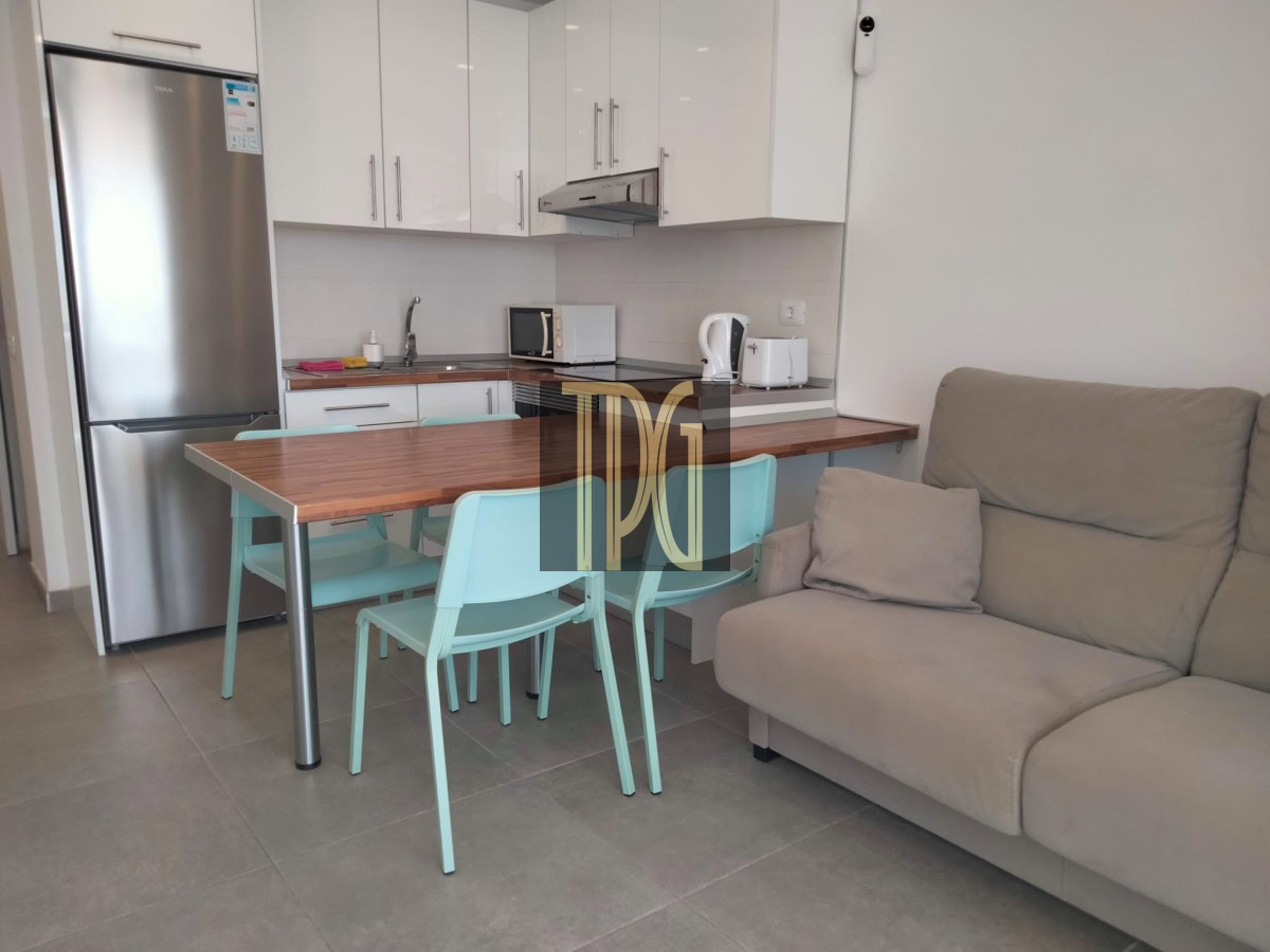 Open-plan kitchen and living area with a wooden dining table and light turquoise chairs beside a beige sofa. Stainless steel refrigerator next to white upper cabinets, with a microwave, kettle, and toaster on the counter. Turquoise dining chairs surround a wooden table in a modern kitchen setup with a gray-tiled floor. Beige sofa in a cozy living space adjacent to a compact, contemporary kitchen in a studio apartment. Modern apartment: kitchen and living area with white cabinets, stainless appliances, and a small dining table.