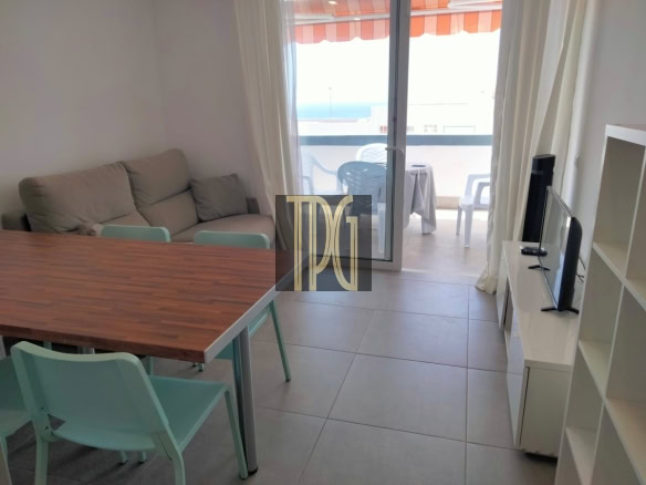Living/dining area with a wooden table, mint chairs, beige sofa, and a TV on a white stand; balcony visible through glass door.