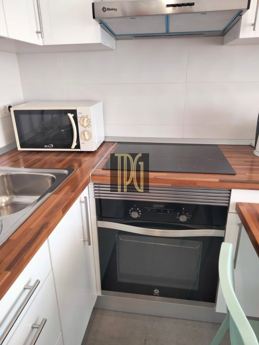 Compact kitchen setup with white cabinets, wood countertops, a microwave on the left, induction hob, and a built-in oven beneath a stainless range hood.
